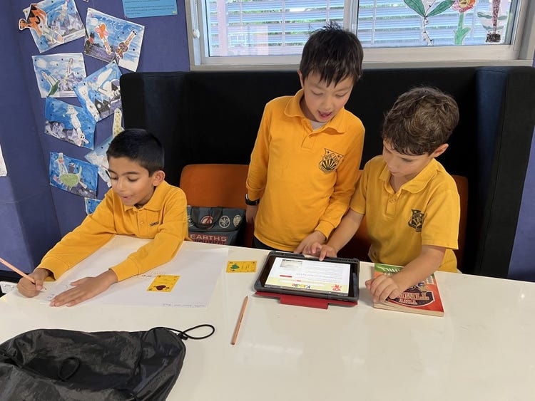 Two students working together on an iPad task while another student works independently nearby.