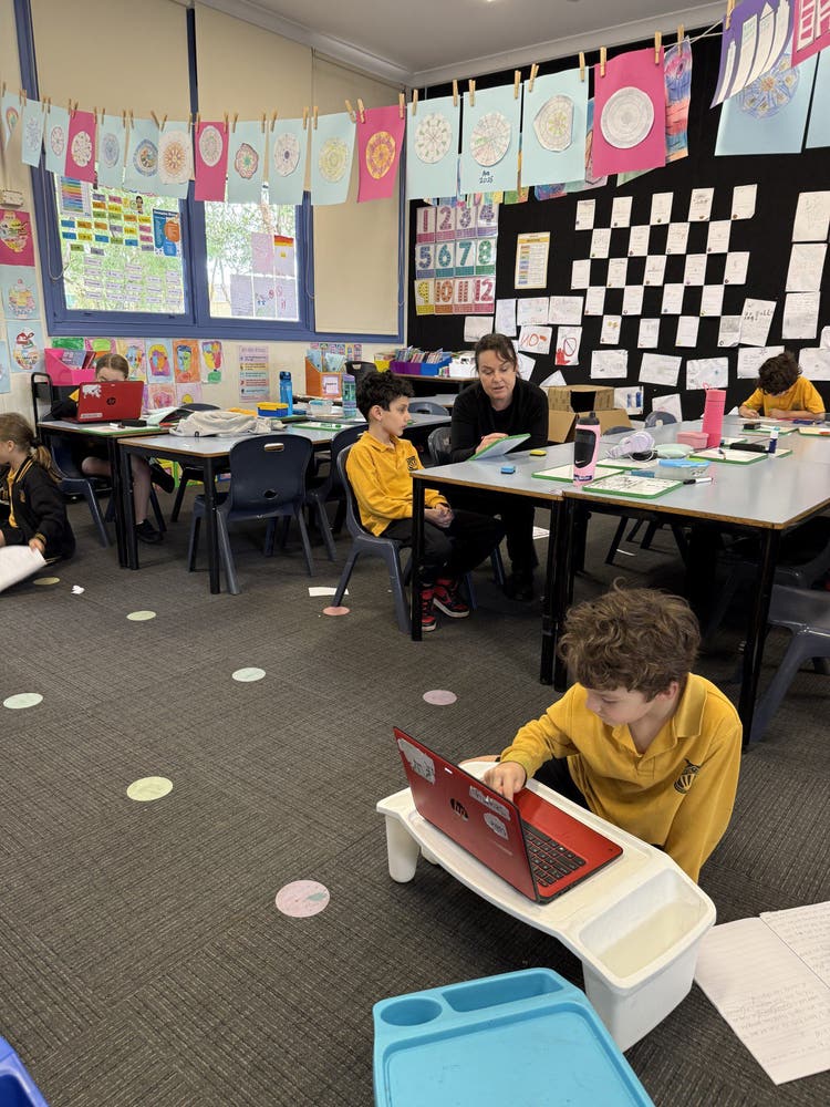 Teacher working one-on-one with a student at a classroom desk, while another student works independently on the mat.