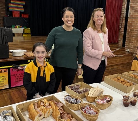 Principal, P&C president, and a student smiling together at a table in the school hall during the sausage sizzle service.
